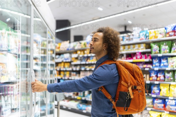 Young african american man with curly hair and a backpack opening a supermarket freezer,