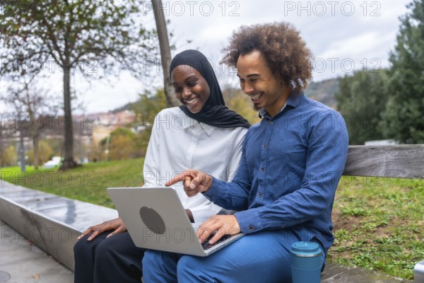 Diverse young couple sitting on a park bench, smiling and pointing at a laptop screen while sharing