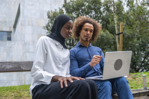 Diverse young man and woman collaborating on a laptop while seated on a park bench, focused on work