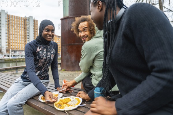 Diverse group of young adults sharing a casual outdoor meal on a city park bench, smiling, talking