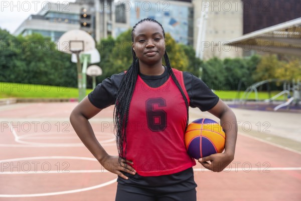 Confident young black woman athlete with braids wearing a red jersey, holding a basketball, and