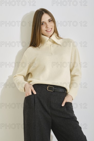 A woman stands by the bright wall with a warm smile, wearing a stylish cream sweater and loose dark