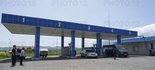 Covered gas station with numbered pillars, several vehicles and people, gas filling station, Sewan,