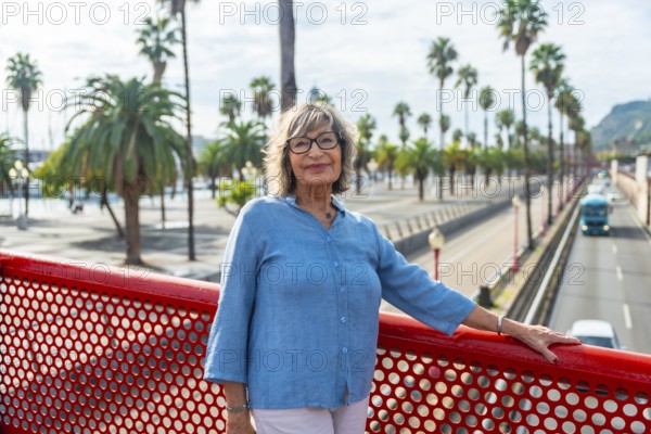 Senior woman standing on a red bridge, smiling directly at the camera, with a vibrant city street
