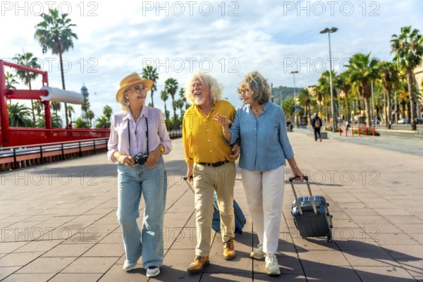 Three cheerful senior tourists walking together on a promenade with palm trees, carrying luggage