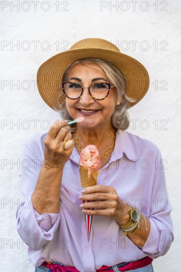 Happy senior woman wearing a straw hat and glasses smiling while eating a refreshing ice cream cone