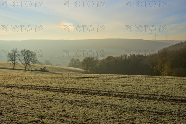 View over a frosty field and distant hills with wind turbines on the horizon at dawn, Seckmauern,