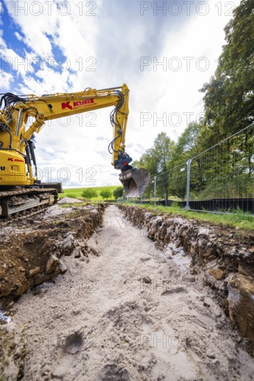A yellow excavator digs a deep ditch surrounded by nature and a cloudy sky, Bau PV