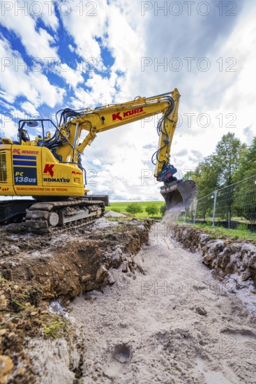 A yellow excavator carries out trenching work on a construction site under a cloudy sky, Bau PV