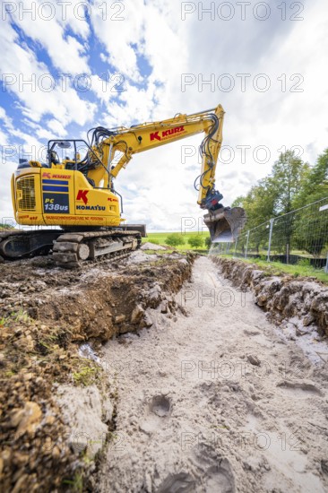 A yellow excavator in a deep ditch surrounded by nature under a cloudy sky, construction PV