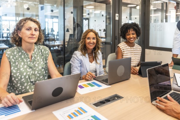 Diverse businesswomen are smiling at the camera, working with laptops and analyzing data graphs