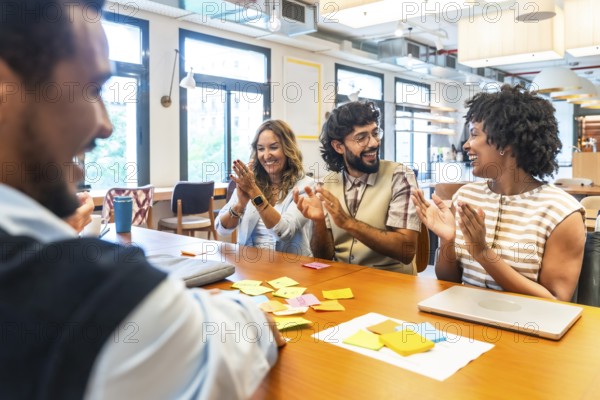Diverse business colleagues sitting at a table in a modern office, happily clapping and smiling