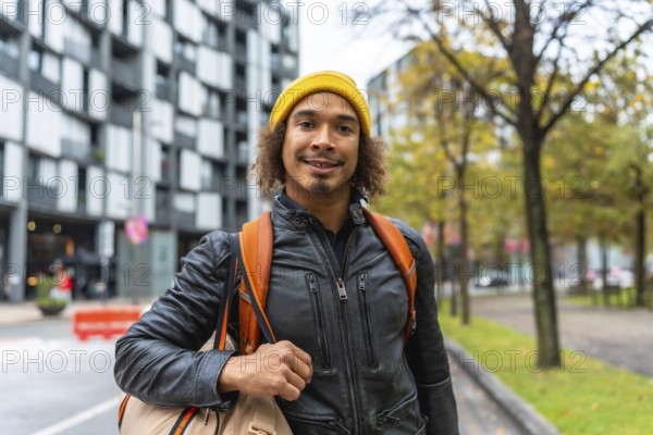 Young man with curly hair in a yellow beanie and leather jacket, backpack on his shoulders, smiling