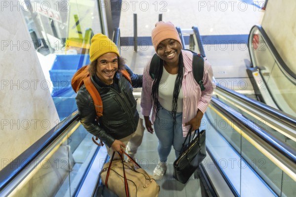 Happy young couple moving up an escalator at an airport or train station, carrying their travel