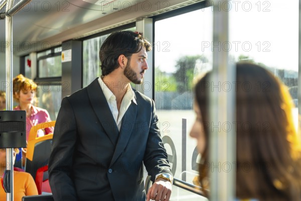Young businessman in a dark suit stands on a city bus, gazing out the window as warm sunlight