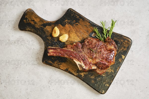 Grilled cowboy steak on a worn wooden cutting board, garnished with fresh rosemary and garlic. The