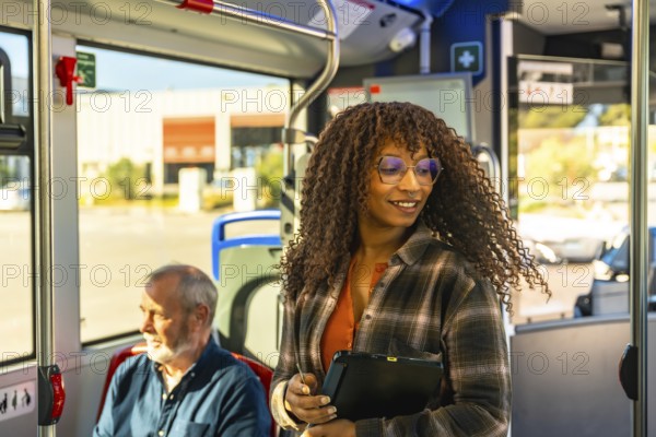 Woman with glasses smiles while standing on a city bus, holding a digital tablet among commuters