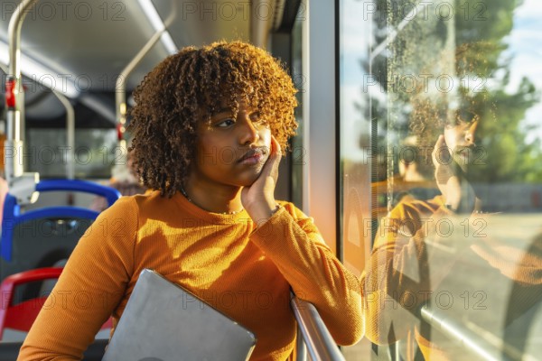 Young woman with curly hair riding public transport bus, holding a laptop and contemplating while