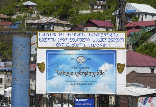 A large advertising sign with a blue poster and text surrounded by colorful buildings and green