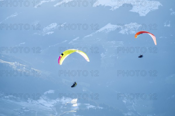 Two paragliders flying in front of snowy mountains in the evening light in winter, Kitzbühel Alps,