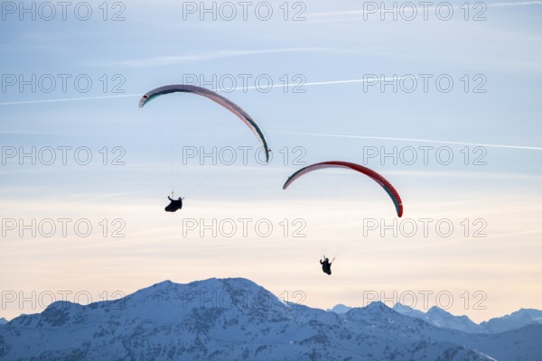 Two paragliders flying in front of snowy mountain peaks in the evening light in winter, Kitzbühel