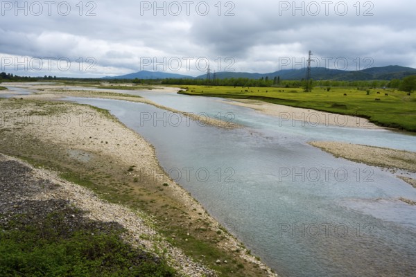 A calm river flows through a pebble landscape under a cloudy sky with power lines in the