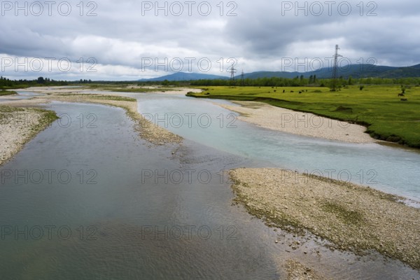 A river snakes through strips of pebbles flanked by green fields and cloudy skies, interrupted by