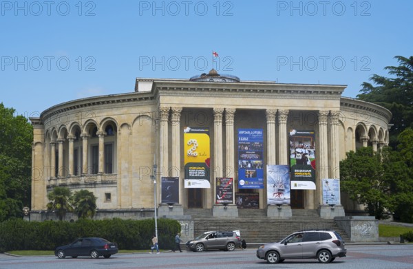 A round classical building with pillars and posters against a blue sky surrounded by green trees