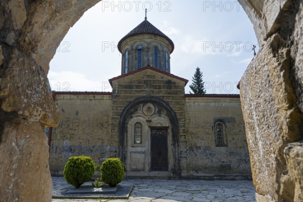 Through a stone arch, you look at an old church entrance and green courtyard, Motsameta monastery,