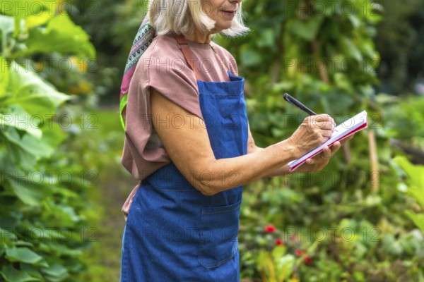 Senior woman in apron and colorful scarf jotting notes in a notepad with a pen while standing in a