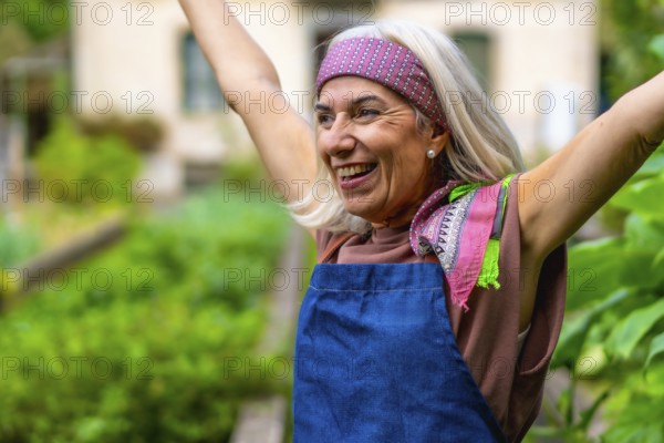 Senior woman in a bandana and apron raises her arms in a sunlit garden, smiling with joy and