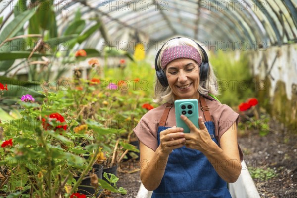 Senior woman wearing headphones and apron, smiling while looking at her smartphone and interacting