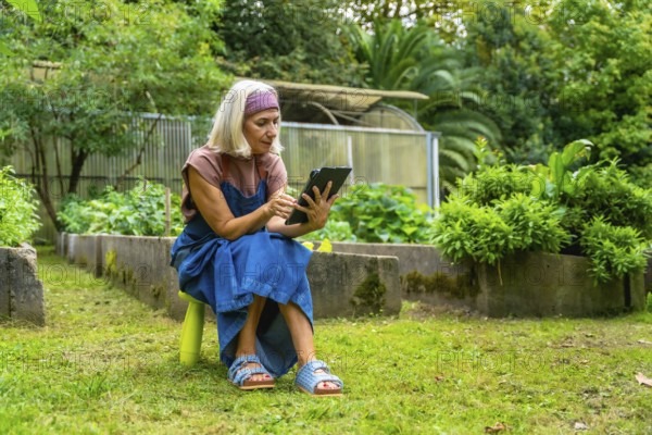 Senior woman sitting on a stool in her green garden, using a digital tablet for managing her plants