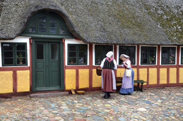 Two women in older farm costumes in front of a half-timbered house with a thatched roof and on