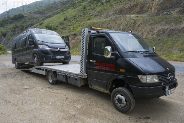 Tow truck transporting van on a rural road with green hills in the background, camper being towed,