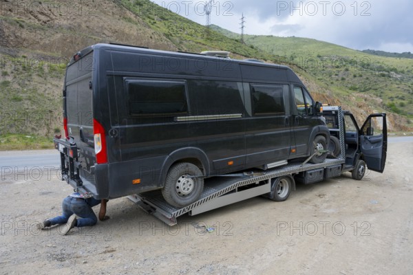 Black van on a car transporter, driver working under the vehicle, camper being towed, motorhome,