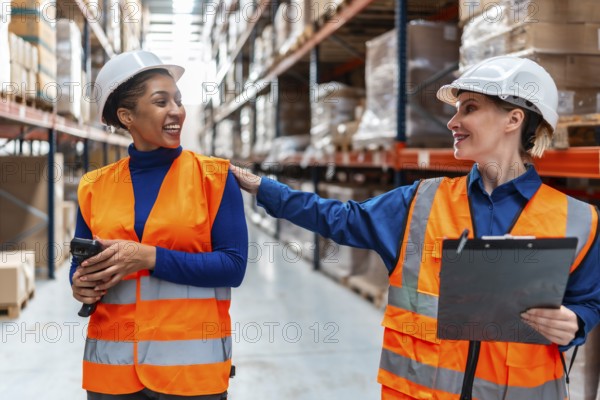 Two diverse women warehouse workers in safety vests and hard hats smiling and collaborating,