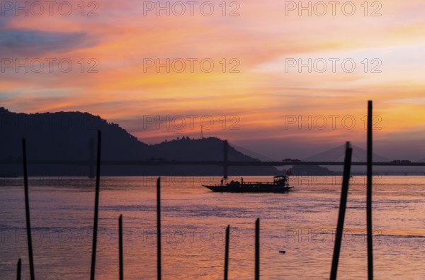 A boat sails on the river Brahmaputra during sunset, with the silhouette of a cable-stayed bridge