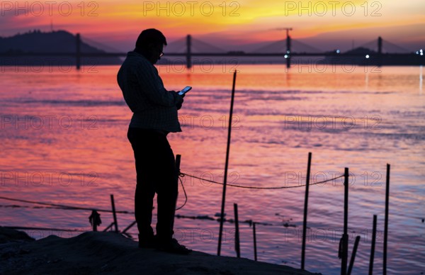 A person browsing his mobile phone by the Brahmaputra riverside, during sunset, on November 4, 2025