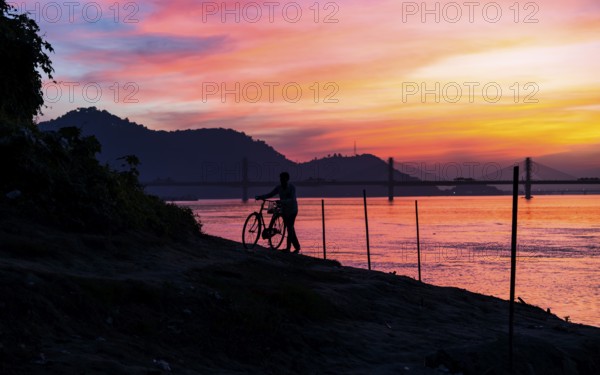A man pushes his bicycle along the Brahmaputra riverbank during a vibrant sunset, with the