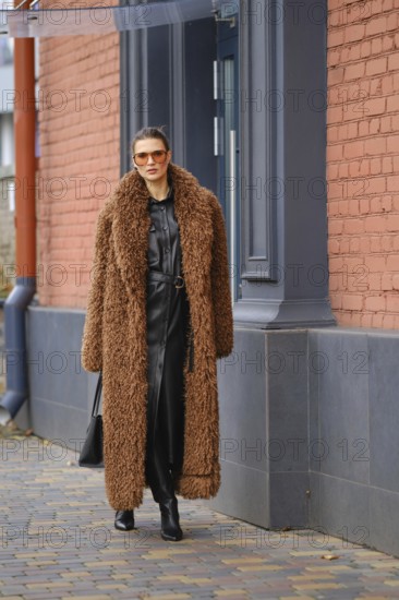 A fashionable woman strolls along a city sidewalk, wearing a cozy brown fur coat. She pairs it with