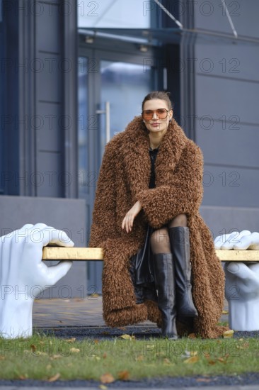 A stylish woman wearing a brown fur coat and black knee-high boots poses on a unique bronze bench