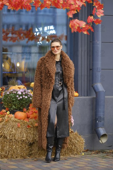A woman dressed in a stylish brown fuzzy coat and black leather dress stands next to a seasonal