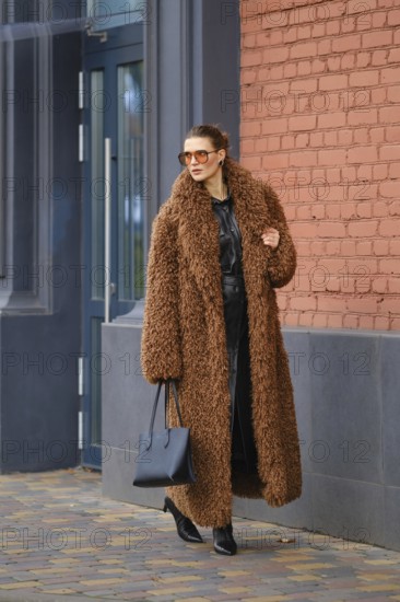 A stylish woman with sunglasses walks along a sidewalk by a vibrant brick wall. She wears a fluffy