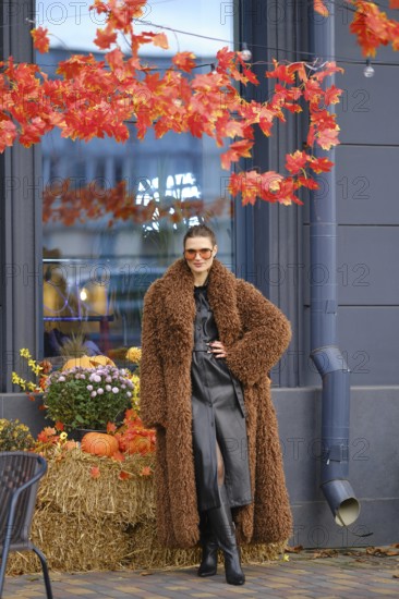 A stylish model stands near a store with bright autumn leaves overhead. She wears a brown fur coat
