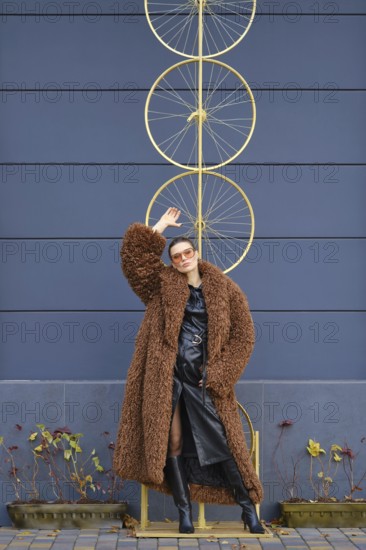 A woman stands confidently near a striking bike wheel art piece. She wears a fluffy brown coat and