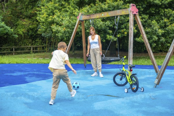 Young boy kicking a soccer ball on a playground while mother watches, with a swing set and bicycle