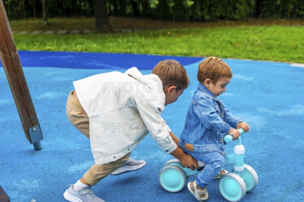 In a vibrant playground setting, an older brother supports his younger sibling as he learns to ride
