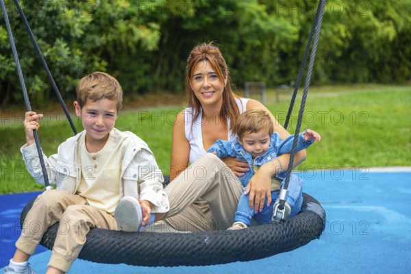 Happy family enjoying a sunny day at the playground, the mother is swinging with her two children
