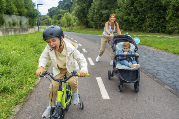 Child wearing a helmet is riding a bicycle and shouting, while his mother is pushing a stroller